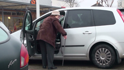 Eine ältere Frau mit Stock steigt auf der Fahrerseite in ein Auto ein.