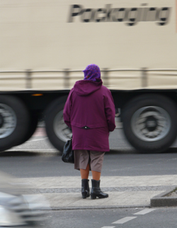 Eine ältere Frau wartet auf einer Mittelinsel darauf, dass ein großer Lkw vorbeifährt