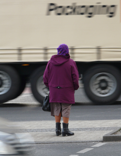 Eine ältere Frau wartet auf einer Mittelinsel darauf, dass ein großer Lkw vorbeifährt Eine ältere Frau wartet auf einer Mittelinsel darauf, dass ein großer Lkw vorbeifährt