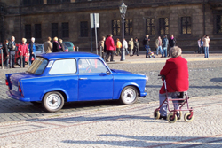 Seniorin sitzt auf einem Rollator an einer schmalen Straße auf der nur ein einziges Auto fährt